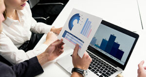 Two professionals reviewing charts and graphs on a printed report while working on a laptop