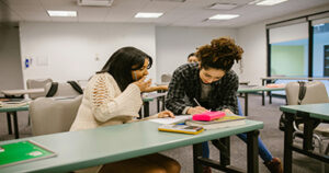 Two students studying together in a classroom while reviewing notes and textbooks.