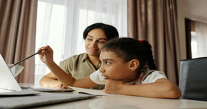 Mother helping her child with homework while using a laptop at home.