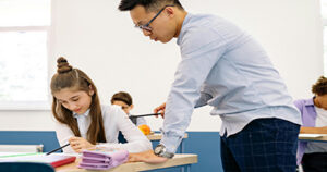 Teacher assisting a student while she works on a classroom assignment at her desk.