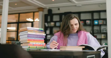 The image shows a woman sitting at a table with a stack of books beside her, reading one of them. She is wearing a purple sweater and appears focused in a well-lit library or study space.