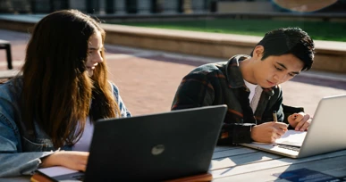 The image shows two students sitting at a table outdoors, one working on a laptop and the other writing in a notebook, with a bright and sunny background.