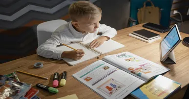 The image shows a young boy sitting at a desk, writing in a notebook while looking at a tablet. The table has books, colored markers, and other stationery items.