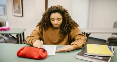 The image shows a young woman sitting at a desk in a classroom, focused on a sheet of paper. There are books and a red pencil case on the table.