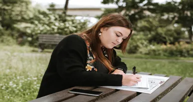The image shows a woman sitting at a wooden table outdoors, writing in a notebook while surrounded by greenery. She appears focused and relaxed.