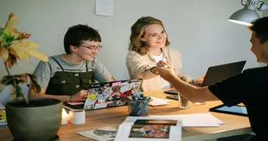 The image shows three people in a casual setting, with two working on laptops and one shaking hands.
