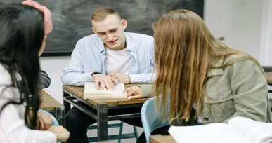 "Three students sitting together at a desk, discussing and studying from a textbook in a classroom setting."