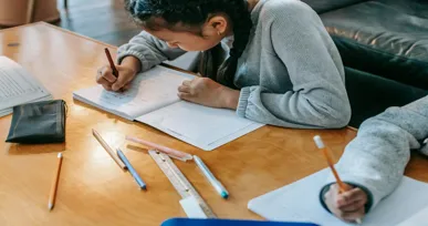 The image shows a child with braided hair, focused on writing in a notebook at a table surrounded by colored pencils and other stationery items