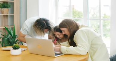 Three students collaborating at a table while working on a laptop and writing notes together.