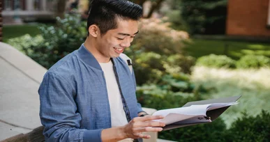 The image shows a young man smiling while looking at a book outdoors, surrounded by greenery in a well-lit environment.
