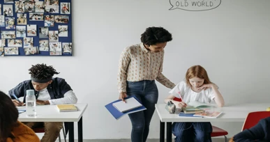 The image shows a teacher standing next to a student at a desk, offering guidance while another student is focused on their work. The classroom has a playful atmosphere with photos on the wall and students engaged in their activities.