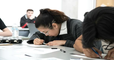 The image shows a student focused on their work, drawing or writing on paper. Other students are visible in the background, working at their desks in a classroom setting.