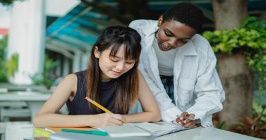 Student writing in a notebook while another student helps and explains the work