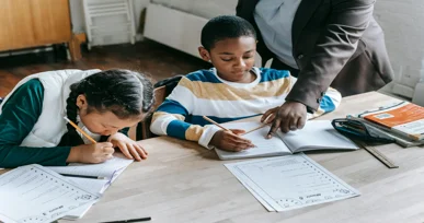 "Two children, one writing in a workbook and the other focused on their studies, with a teacher assisting them at a desk in a classroom setting."