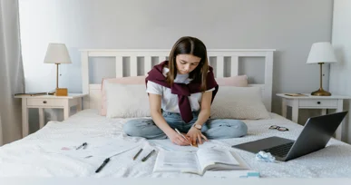 "A student studying on a bed with an open textbook, laptop, and stationery, focused on writing in a relaxed and cozy room."