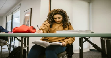 A focused student sitting at a table, studying and writing in a textbook in a bright, modern study room.