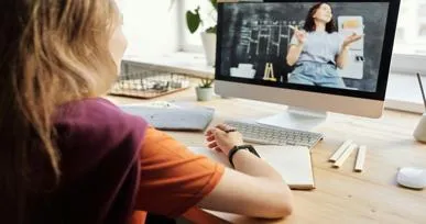 The image shows a person sitting at a desk, looking at a computer screen displaying a woman in a virtual meeting or lesson. The background has a bright, airy atmosphere with plants and a clean desk setup.
