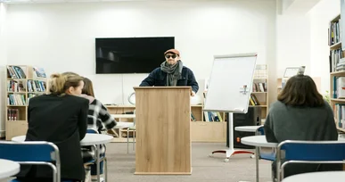 The image shows a person standing at a podium in front of a classroom with students sitting at desks. The person is wearing sunglasses and a scarf, while the students are facing the speaker in a library-like setting.
