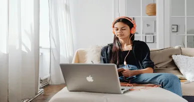 The image shows a young woman sitting on a couch, wearing headphones and focused on her laptop, with a relaxed and well-lit environment.
