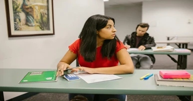 "A student sitting at a table in a classroom, looking to the side while studying, with notebooks and stationery on the desk, and a classmate working in the background."