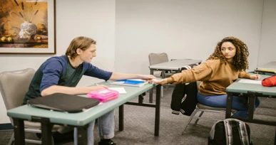 "Two students sitting at desks in a classroom, reaching across the table to exchange notes or materials, with books and stationery on the desks."